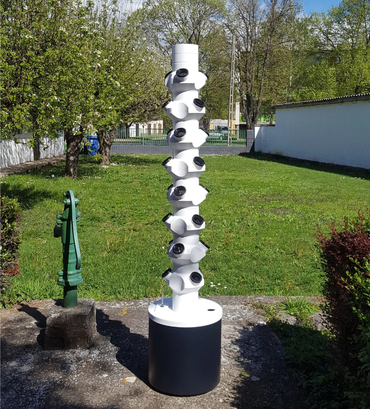 Weißer Turm mit Pflanzen im Garten, davor eine grüne Handpumpe, dahinter blühende Bäume